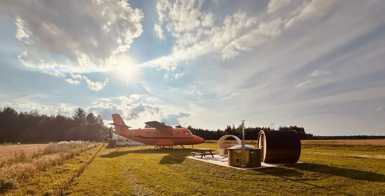 Oranžové letadlo na louce u lesa, vedle sauna a koupací sud, zlatá hodina